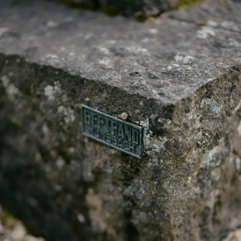 Plaque en bronze Bertrand Münsbach sur un socle en pierre couvert de lichen, gravier clair en contrebas